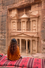 A girl in an orange shirt sits on the edge of a cliff on a carpet and looks at the ancient city of...