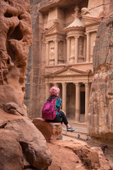 Girl tourist with a backpack sits on a stone and looks at El-khazneh in the Siq canyon - obrazy, fototapety, plakaty
