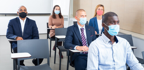 Fototapeta na wymiar International group of business people wearing protective face masks listening to presentation in conference room. Concept of precautions and social distancing in COVID 19 pandemic..