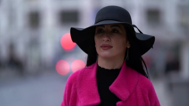 Close-up portrait of a stylish black-haired woman in a pink coat and black hat walking and posing on a blurry background of a busy city street.