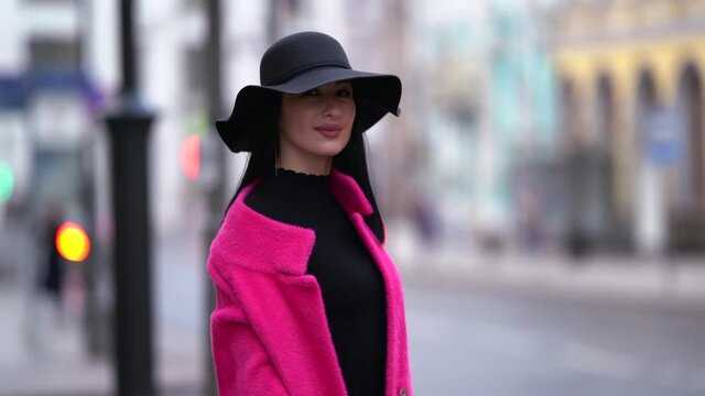 A stylish black-haired woman in a pink coat and black hat walks and poses on a blurry background of a city street near a highway.