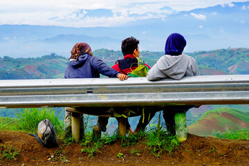 Three young people sit overlooking a valley of fields in a village leaning against metal street separator. 