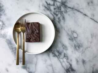 Brownies on a white plate are served with golden spoons on a marble table.