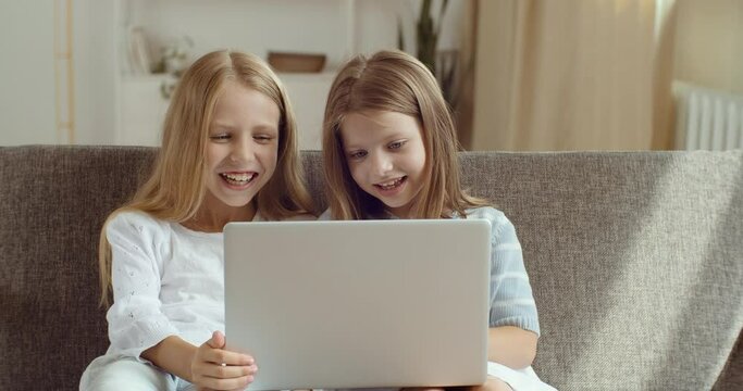 Direct view of two little smiling sisters looking at laptop. Portrait of girls children use computer to watch cartoons movies funny videos in Internet online laughing together sitting on couch at home