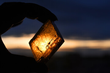 A silhouette image of a hand holding a piece of honey calcite crystal against a golden sunset sky. 