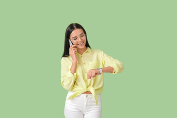 Young woman with wristwatch talking by phone on color background