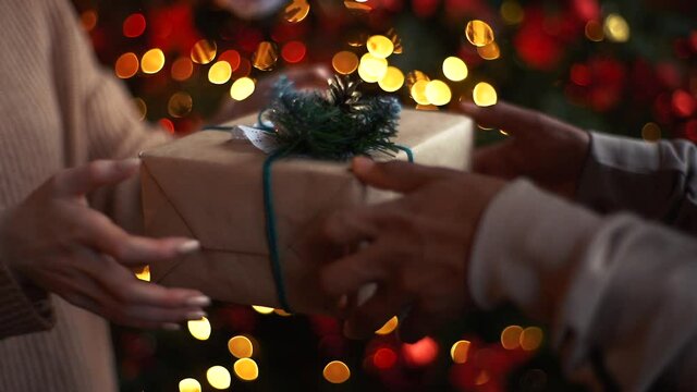 Close-up hands of Caucasian young woman giving Christmas gift to African American man on background Christmas tree with bright blurred lighting at cozy dark living room, slow motion, side view.