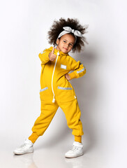 Self-confident cute dark-skinned kid girl in warm yellow trendy jumpsuit and white sneakers stands...