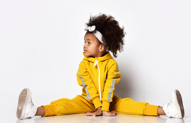 Cute dark-skinned kid girl in warm yellow sport jumpsuit and sneakers sits onm floor with legs spread wide