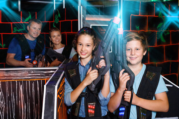 Happy glad cheerful teen brother and sister standing with laser pistols during laser tag game with parents in labyrinth