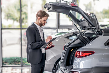 Bearded male examining open car boot, taking notes in the showroom