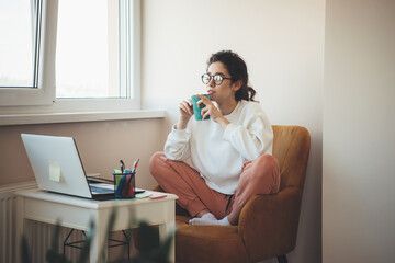Photo of a concentrated woman with glasses drinking tea at home sitting in an armchair and watching...