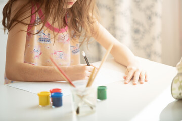 child in pajamas on a sunny morning draws with a pencil on paper at a white table. There are cans of paint and brushes on the table