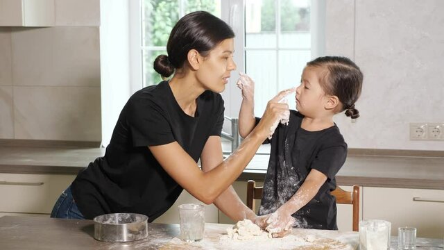 Lovely daughter and mom wearing same black shirts and fancy hairstyle play with flour and dough in beautiful white modern kitchen