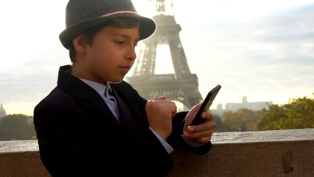 A happy teenage boy in a grey hat is clicking on the phone screen on the background of the Eiffel tower at the sunrise, Paris, France