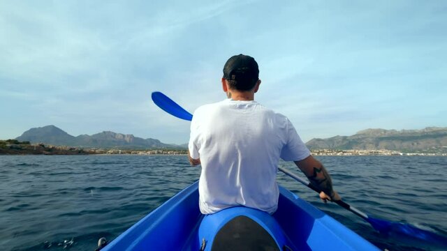 Rear view of man in white tshirt paddle in open waters inside blue kayak. Active healthy lifestyle in great outdoors. Sporty modern man on active weekend 