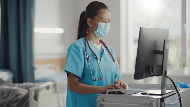 Hospital Ward: Professional Experienced Chinese Head Nurse / Doctor Wearing Face Mask Uses Medical Touch Screen Computer, Checking Patient's Medical Data. In The Background Patient Recovering On Bed 