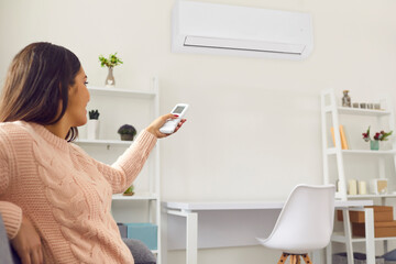 Happy woman sitting on sofa in living-room and switching on air conditioner with remote control
