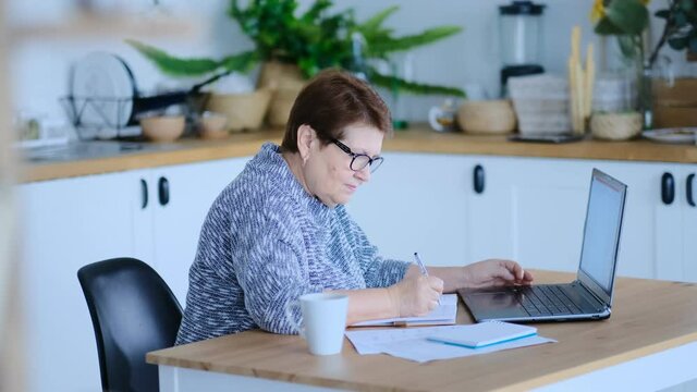 Senior woman using laptop for websurfing in her kitchen. The concept of senior employment, social security. Mature lady sitting at work typing a notebook computer in an home office. Shot video