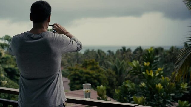 Young athletic man stands on the balcony of house, looks dramatic rain clouds over the sea and filming a movie on smartphone