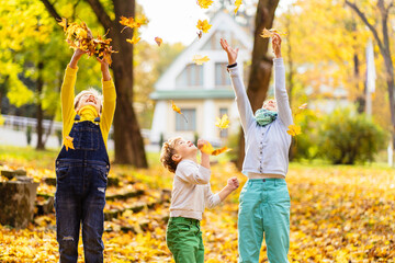 Three children in park playing with fallen leaves. Delighted happy school kids friends having fun throwing fallen leaves up in autumn park.