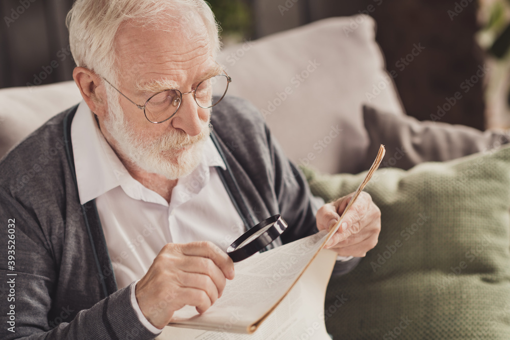 Poster Photo of handsome pensioner wear grey cardigan glasses sitting couch reading newspaper holding loupe indoors house flat