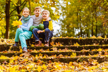 Three children sitting on old leafy stairs in autumn park.