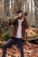 Caucasian boy in beautiful forest with red leaves taking a picture with professional reflex camera photography student with brown jacket, white t-shirt and red scarf, it's cold