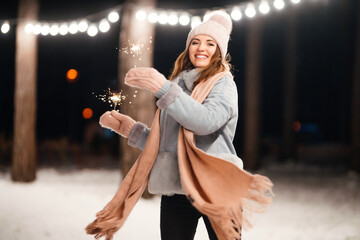 Cheerful young woman celebrating holding sparkles in the winter forest. Festive garland lights. Christmas, new year.