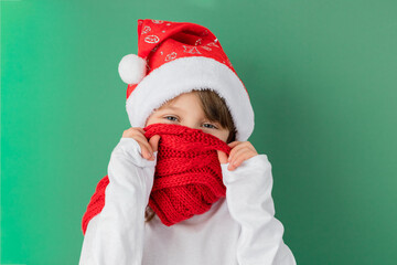 Happy new year and merry christmas. Cute smiling caucasian girl wearing a santa hat covers her face with a red scarf on a green background. Holidays fun. Copy space