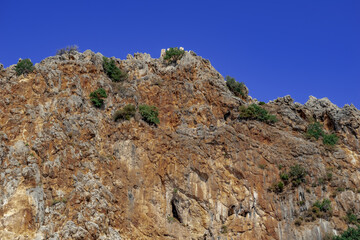 Stone cliff with the ruins of the walls of the Alanya Castle (Turkey) at the top. Brown-orange rock with green trees against blue sky. Natural background with copy space