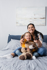 excited girl pointing with finger while watching movie with mother and teddy bear