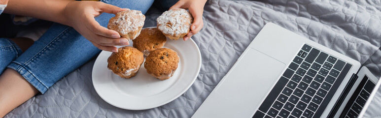 partial view of mother and daughter taking muffins from plate while watching movie on laptop, banner
