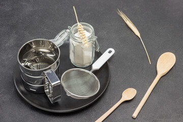 Mug for sifting flour and small sieve,  glass jar with flour on black plate. Wheat spikelets and wooden spoon
