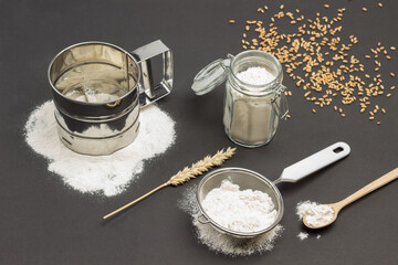 Mug for sifting flour and small sieve with flour. Wheat spikelet and wooden spoon. Wheat grains on table.