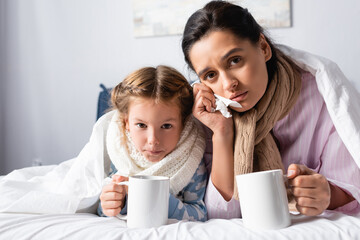 sick, upset woman and daughter looking at camera while lying in bed with warm drink