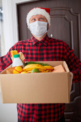 Volunteer man in Santa hat and protective mask and gloves delivery donation box at home in Christmas