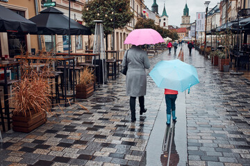 Back view of mother and her daughter holding the pink and blue umbrellas walking in a downtown on rainy gloomy autumn day