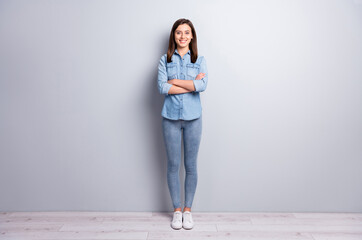 Full size photo of optimistic nice brunette hairdo girl stand crossed arms wear blue shirt jeans sneakers isolated concrete grey wall background