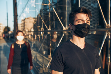 Young man walking in the city center along glass store front in the evening, looking away, wearing the face mask to avoid virus infection and to prevent the spread of disease in time of coronavirus