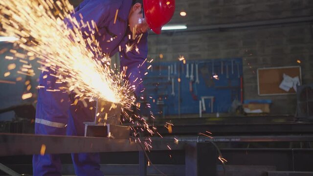 asian male technician worker in a safty uniform wear welders leathers,Electric wheel grinding on steel structure in factory workshop in the garage, orange bokeh sparks fly to the sides