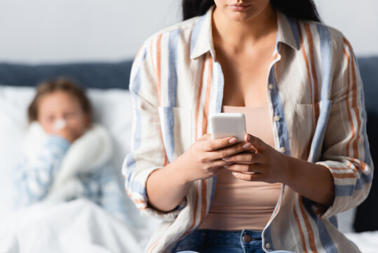 woman messaging on mobile phone near diseased child lying in bed on blurred background