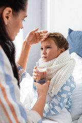woman giving glass of water to ill daughter while touching her forehead on blurred foreground