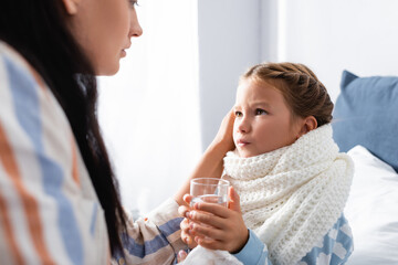 sick girl holding glass of water and looking at mother on blurred foreground