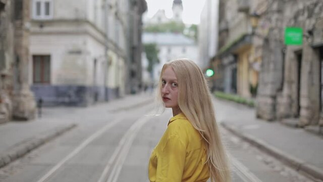 Backside view of young woman with long blond hair walking at old city street. Beautiful female person turning head and looking to camera while keep going. Concept of follow me.