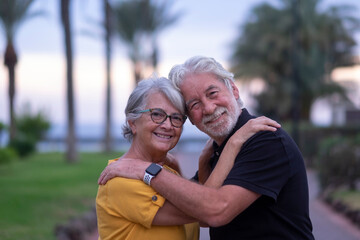 Portrait of two white-haired senior people smiling happily during retirement. Standing outdoors in a public park hugging each other while looking at camera. Serene and in love couple