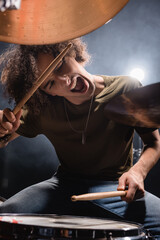 Curly musician shouting while playing drum with drumsticks on blurred foreground