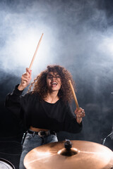 Female drummer with closed eyes shouting, while playing on drum plate with smoke on black background