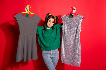 Photo of young attractive doubtful unsure female hold two dresses can't choose isolated on red...