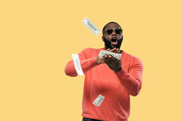 Crazy happy african american guy in sunglasses throws money standing on isolated yellow background